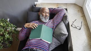 A man in a striped sweatshirt takes a nap on the couch after reading a book