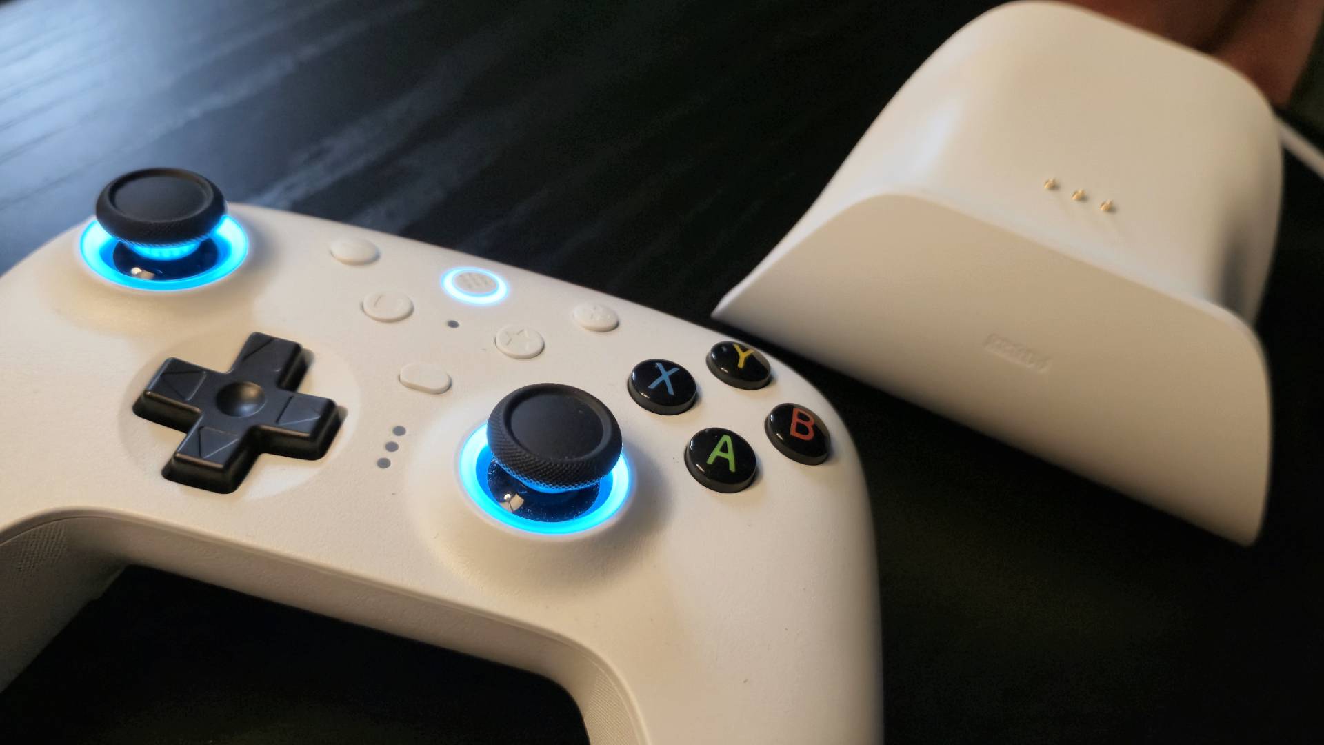 Image of a white wireless controller sitting on a black wooden table, with its charging stand sitting next to it.