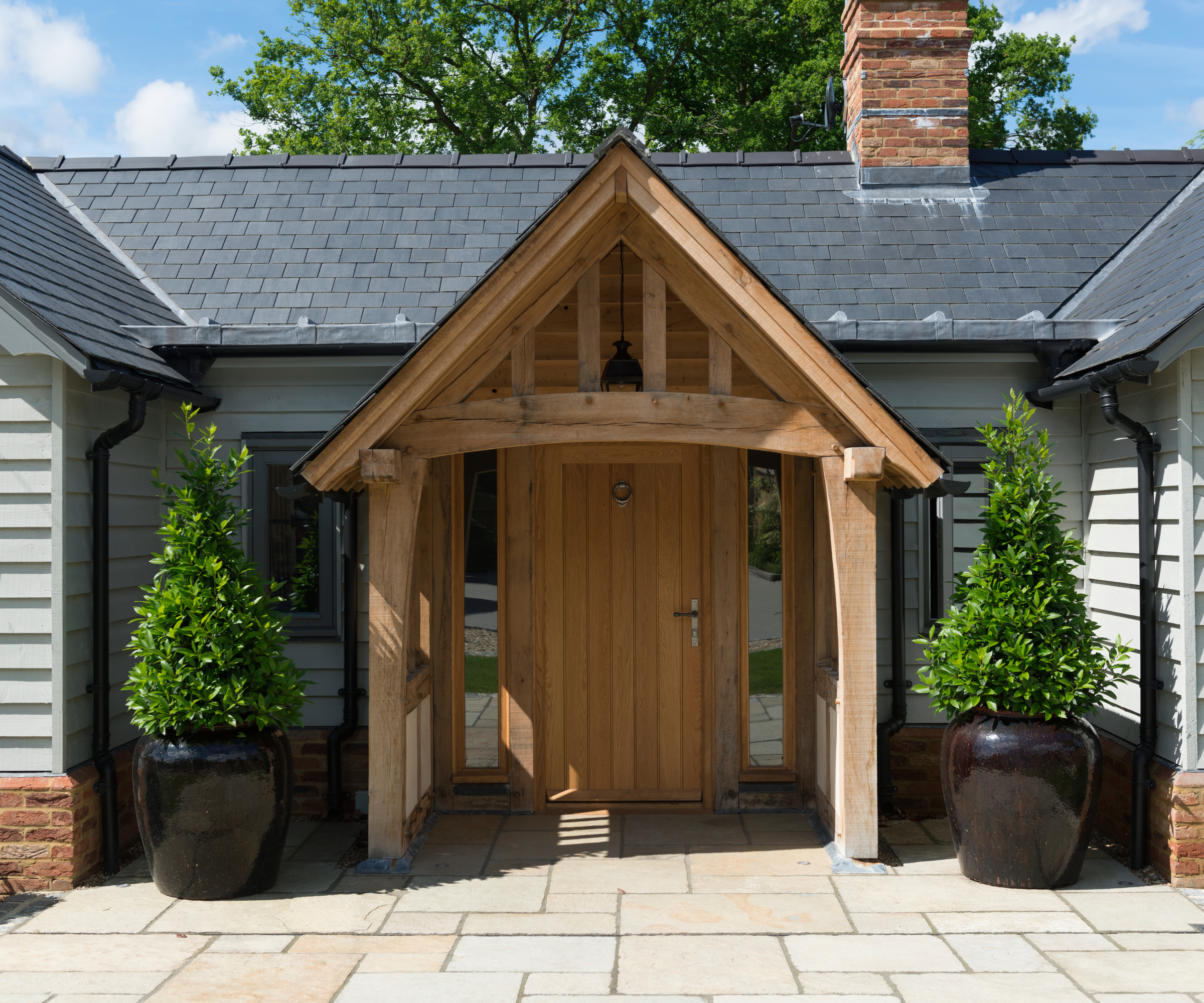 large oak frame porch with pitched roof on single storey home with light grey cladding black tiled roof and front paved entrance leading to oak door