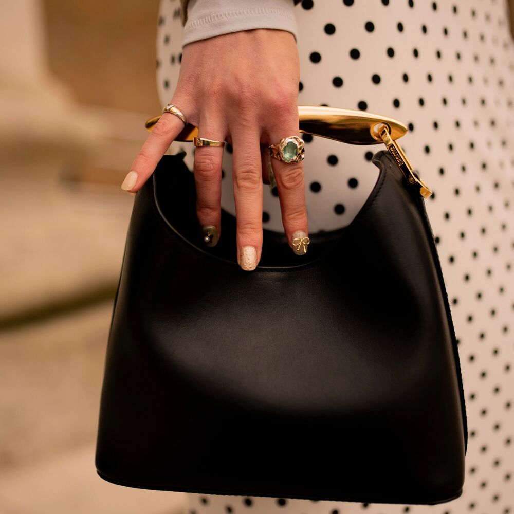 pictures is a a hand with a few rings and a pretty manicure holding onto the handle of a black handbag