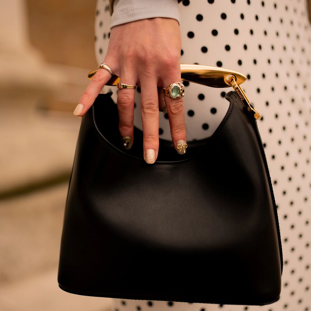 pictures is a a hand with a few rings and a pretty manicure holding onto the handle of a black handbag
