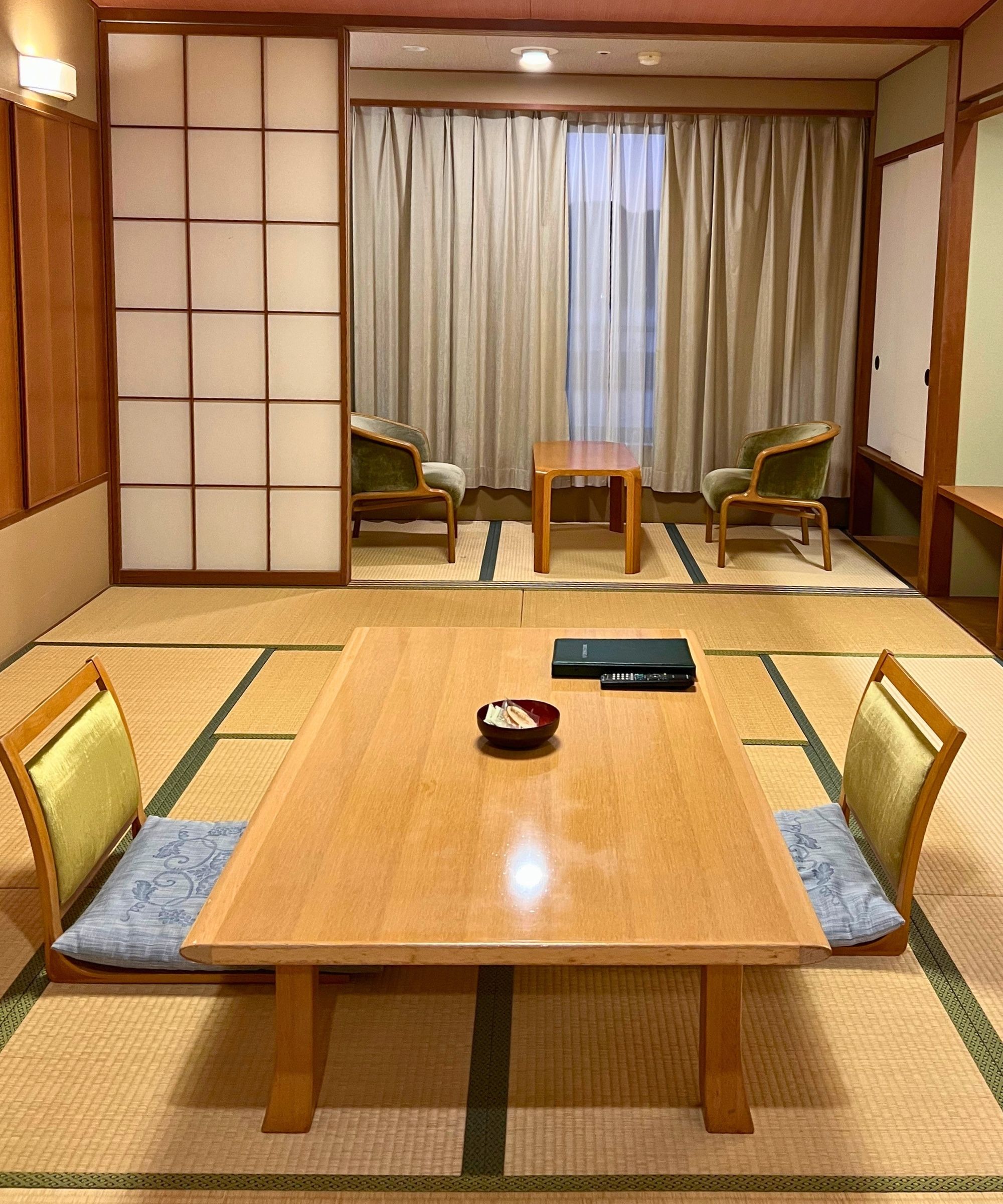 Japanese hotel room with tatami mat flooring, a low seating area, and another seating area in front of large windows with sheer curtains.