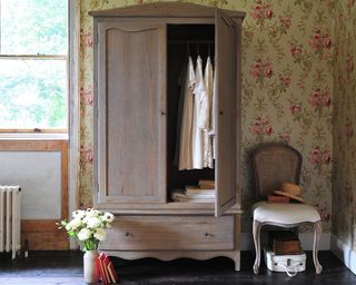 A limewash wooden wardrobe with wooden chair and patterned floral wallpaper in bedroom