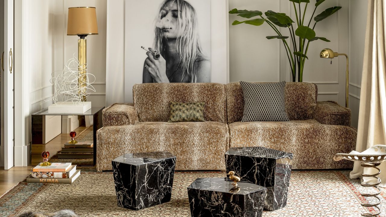 Image of a white living room that is decorated in an eclectic style. There is a cheetah print sofa with three black marble coffee tables in front of it in varying sizes. There is a mirrored side table beside the sofa and a gold floor lamp behind it. There is a photo of a woman and a large floor plant on the back wall.