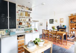 a freestanding kitchen with lots of wooden units, and a wide open white shelving unit on the wall full of jars, a tiled floor and an orange rug under a wooden dining table