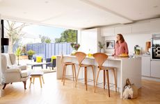 An open plan kitchen with white marble-effect island, wood bar stools, bi fold doors out onto a garden deck, parquet flooring
