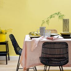 Dining room with yellow wall and table covered with light pink tablecloth and black rattan dining chairs