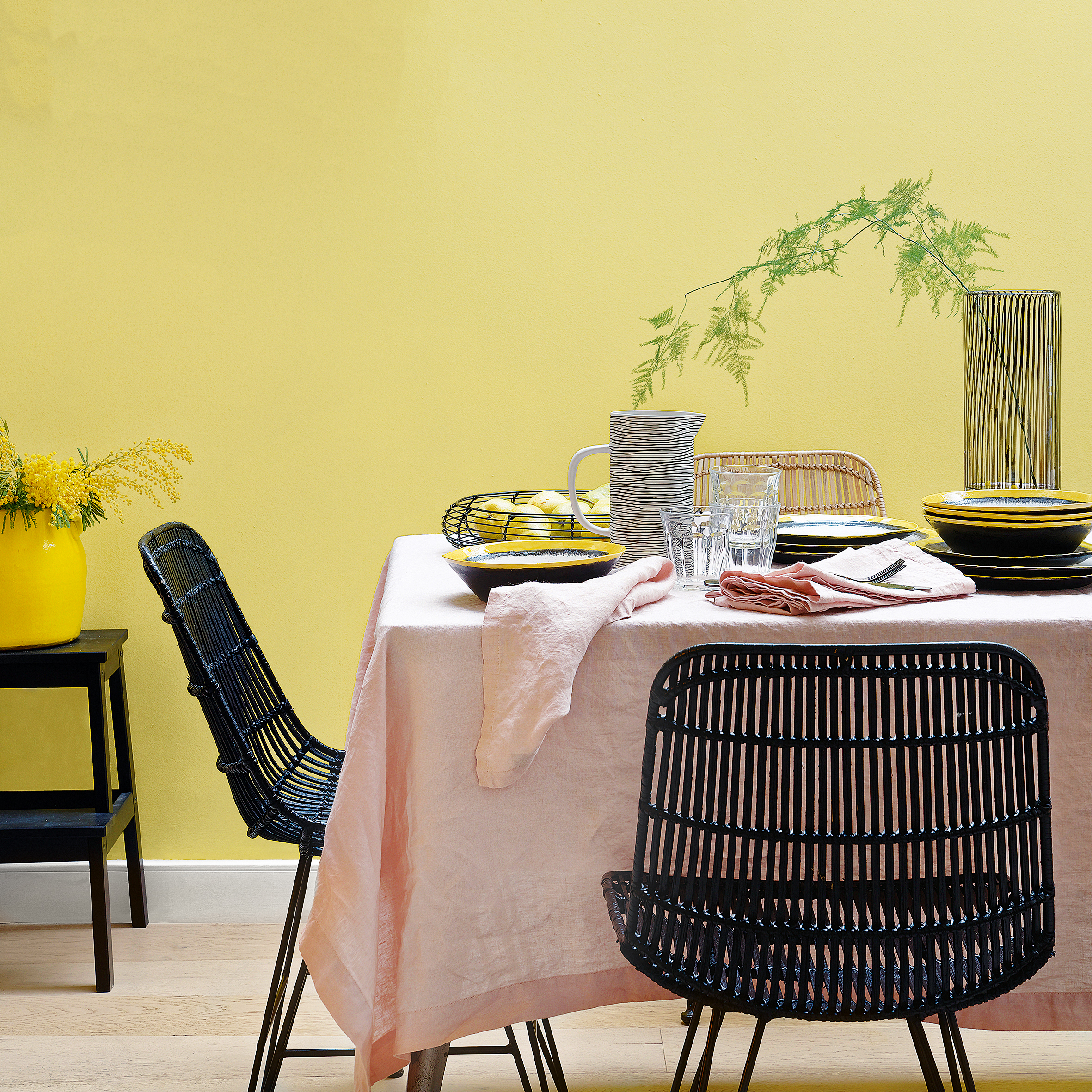 Dining room with yellow wall and table covered with light pink tablecloth and black rattan dining chairs