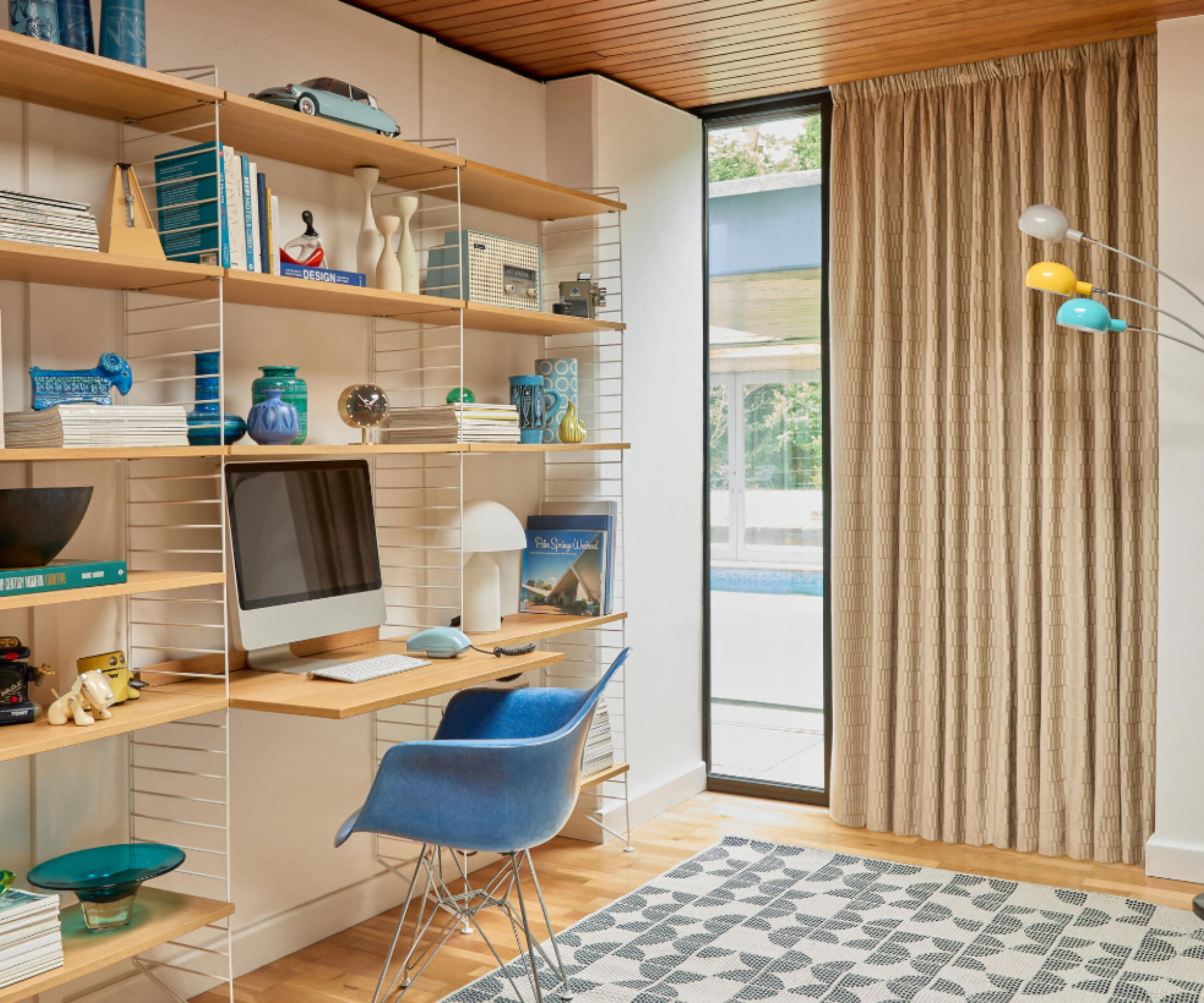 Bright mid-century style home office with floating wooden shelves, a blue chair, retro accessories, and a colourful floor lamp beside tall curtains and a window.