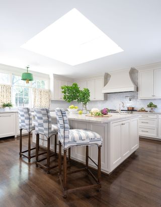 kitchen with skylight over pale island and three blue and white upholstered bar stools green pendant light and patterned curtains