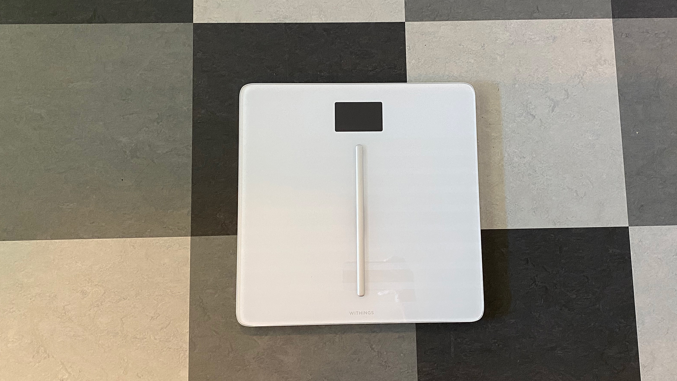 En vit smartvåg av typen Withings Body Cardio står på ett rutigt golv.