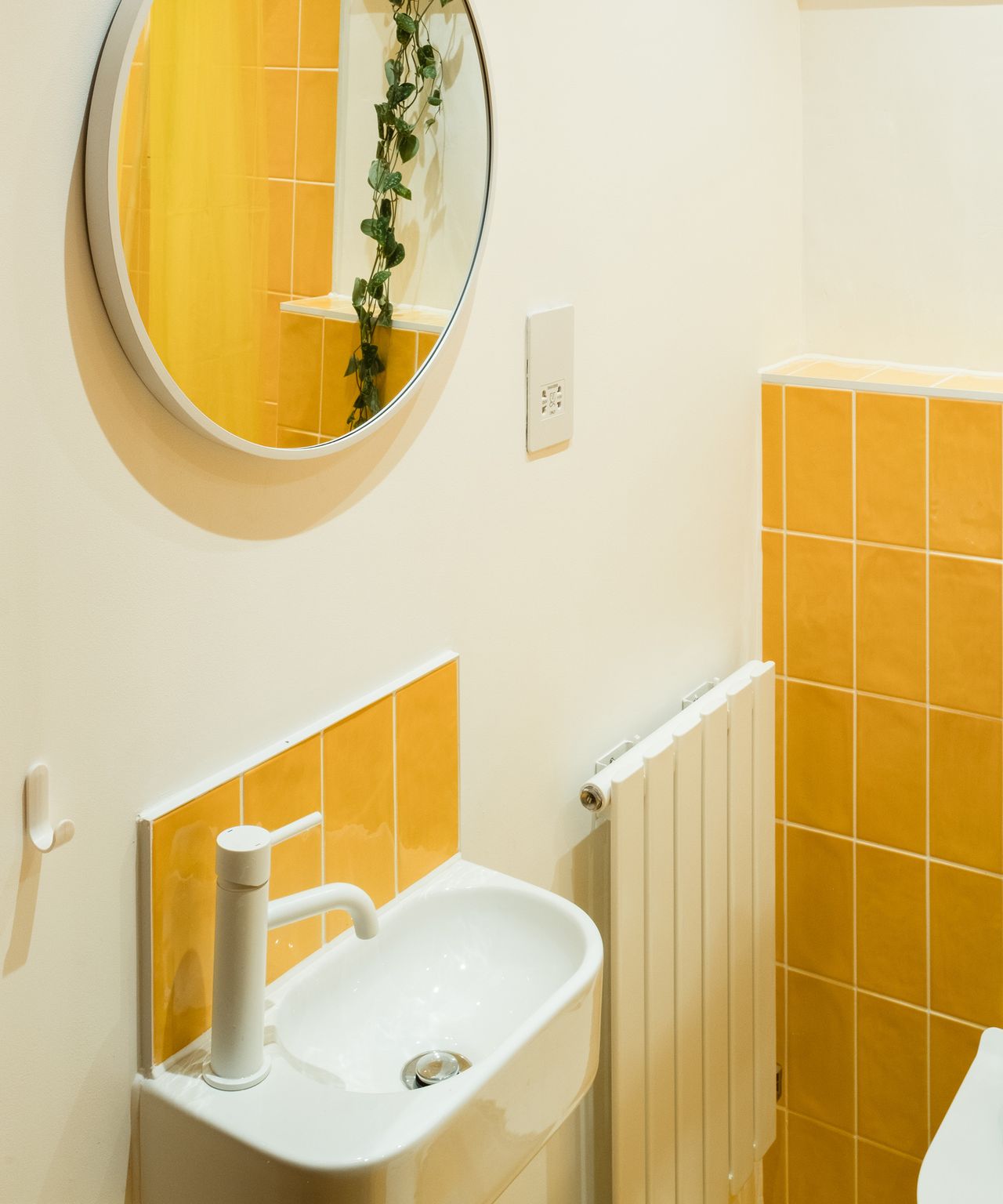 A small bathroom sing with white taps and yellow tiles as the backsplash and behind the toilet. There is a round mirror above the sink reflecting a plant