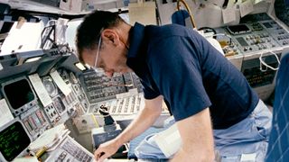 a man in a short-sleeved blue shirt operates a computer on the space shuttle