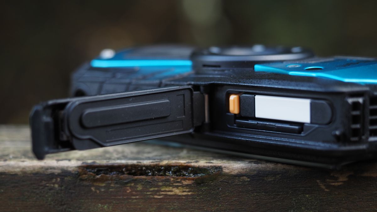Close up of the battery compartment of a Pentax WG-90, sitting on a wooden surface in front of woodland
