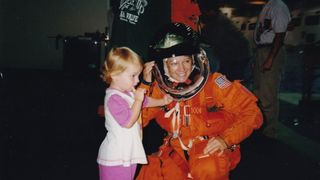 a smiling woman in an orange flight suit kneels next to a young girl