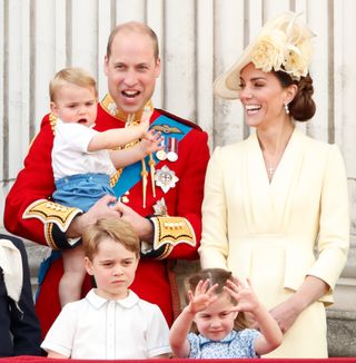 Prince William wearing a red military uniform holding baby Prince Louis and laughing next to Kate Middleton, wearing a yellow dress with a matching hat, and Prince George and Princess Charlotte on the balcony of Buckingham Palace