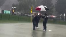 Hideki Matsuyama working on his putting on a flooded green with a team member holding an umbrella over him
