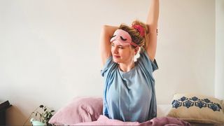 A blonde woman wearing a blue t-shirt and pink eye mask on her forehead with messy bun sitting in bed as she stretches her arms overhead, waking up early to join the 5am club