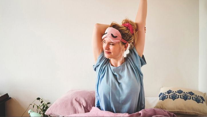 A blonde woman wearing a blue t-shirt and pink eye mask on her forehead with messy bun sitting in bed as she stretches her arms overhead, waking up early to join the 5am club