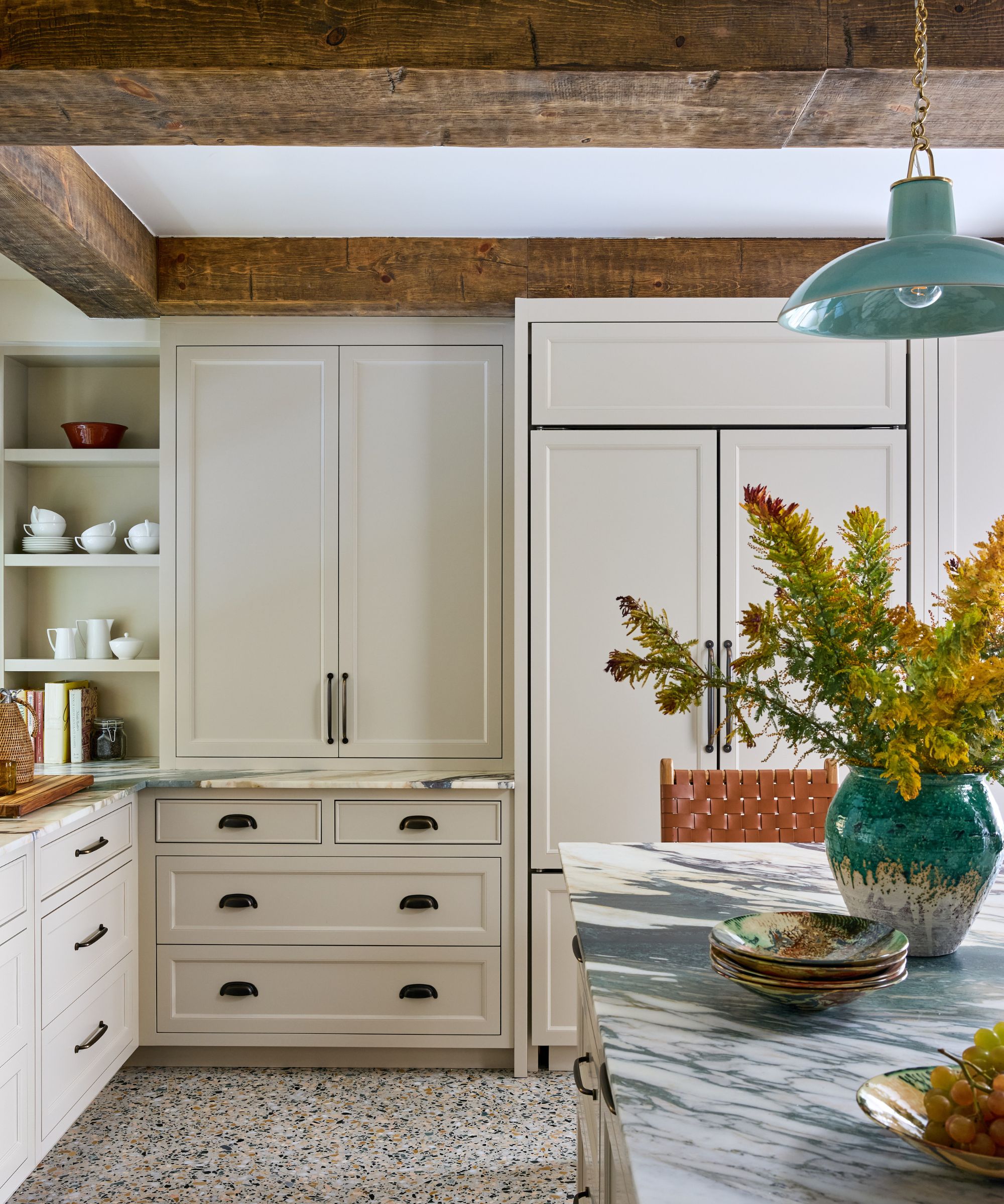 A kitchen with full-height neutral cabinets, wood ceiling beams, terrazzo flooring, and a marble kitchen island with a vase of yellow flowers.