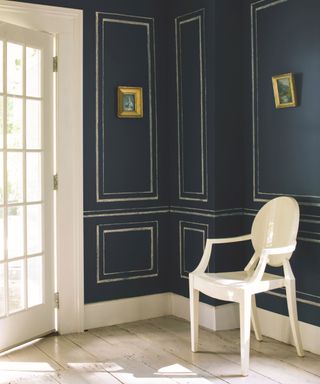 Hallway painted navy blue, gold painted paneling effect, two gold picture frames on walls, white chair and light wooden floor