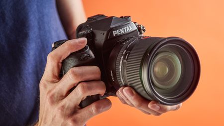 Pentax K-1 DSLR being held in someone's hands in studio with orange backdrop