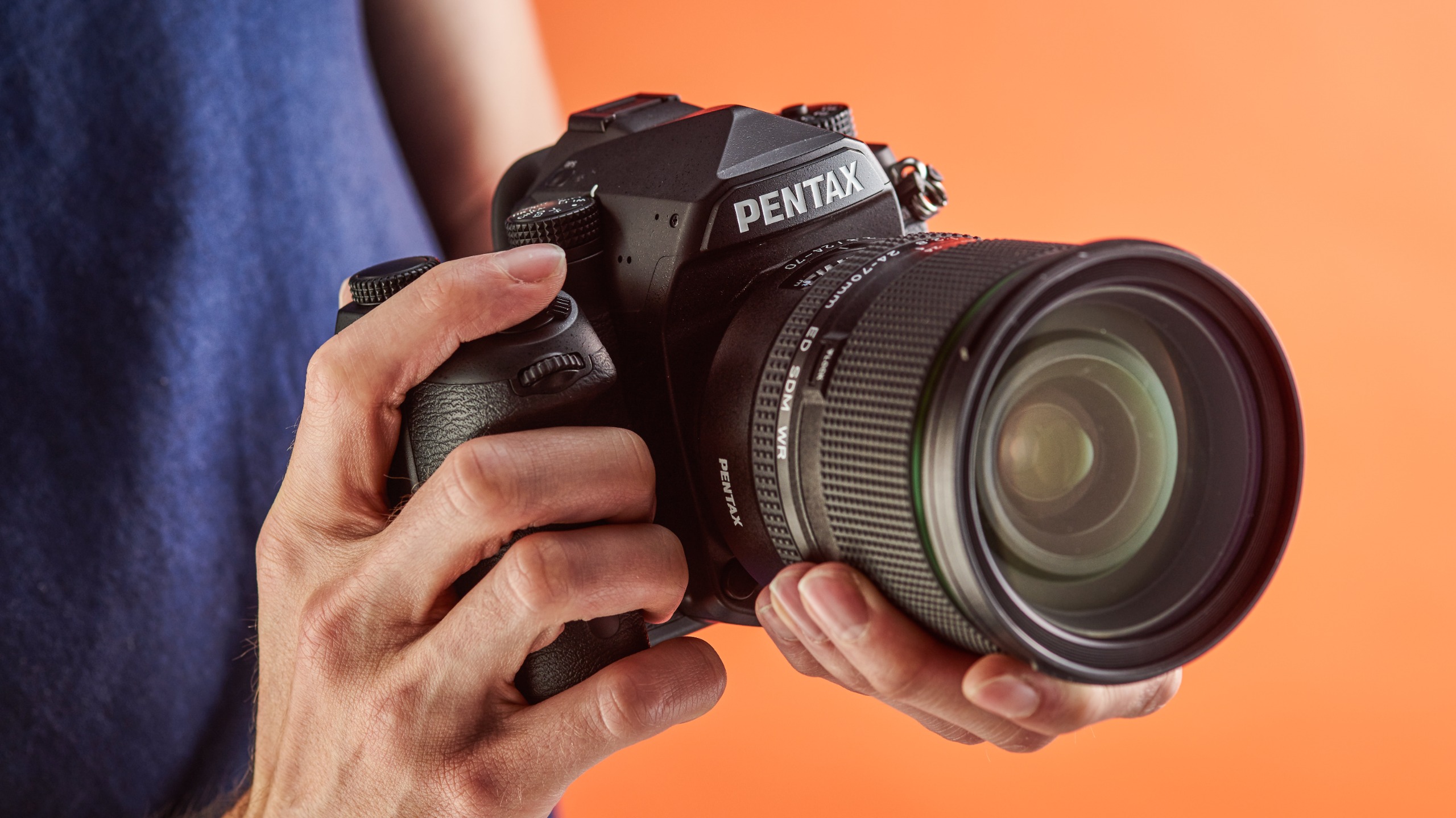 Pentax K-1 DSLR being held in someone's hands in studio with orange backdrop