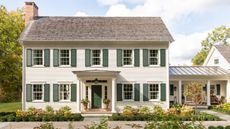 A large traditional house exterior with warm white siding, a dark green front door and dark green shutters, with a porch and flower beds at the front of the home. 
