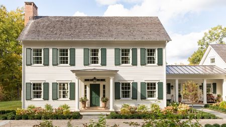 A large traditional house exterior with warm white siding, a dark green front door and dark green shutters, with a porch and flower beds at the front of the home. 