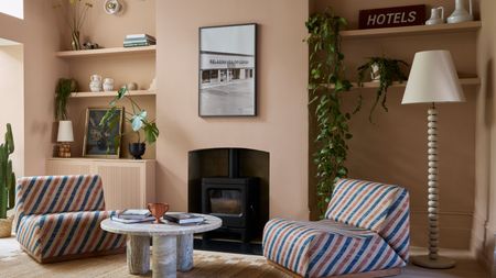 A modern living room color-drenched plaster pink with a log burner, two alcoves either side with open shelving and decorative objects, two red and blue striped low-form chairs, wood floors, and a modern, round coffee table. 