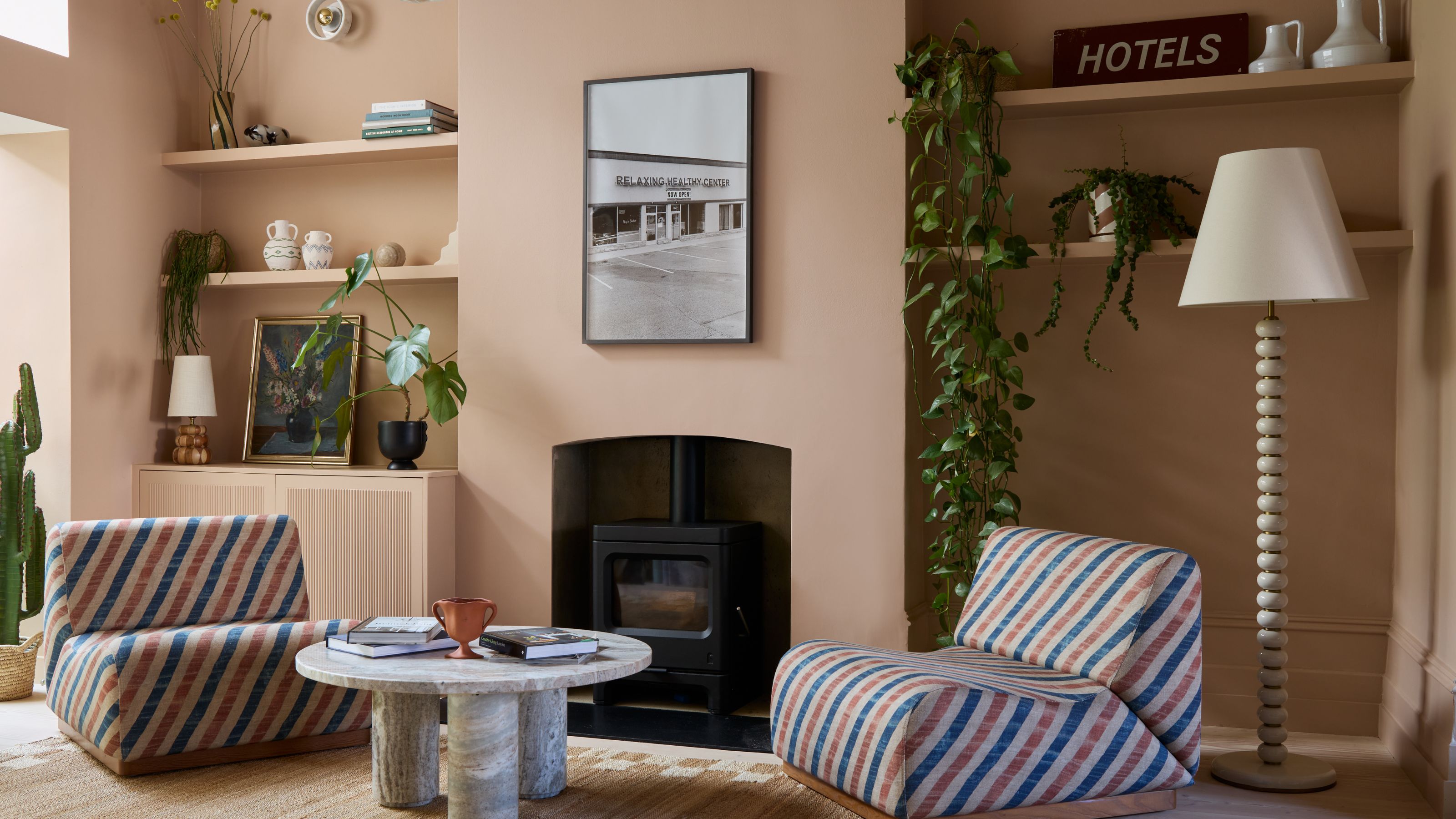 A modern living room color-drenched plaster pink with a log burner, two alcoves either side with open shelving and decorative objects, red and blue striped chairs, wood floors, and round coffee table 