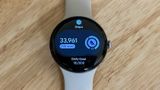 A photo of the Google Pixel Watch 4 on a light wooden desk showing the "Steps" Tile, showing a current count of 33,961 with "23k over" and a Daily Goal of 10,000 underneath.