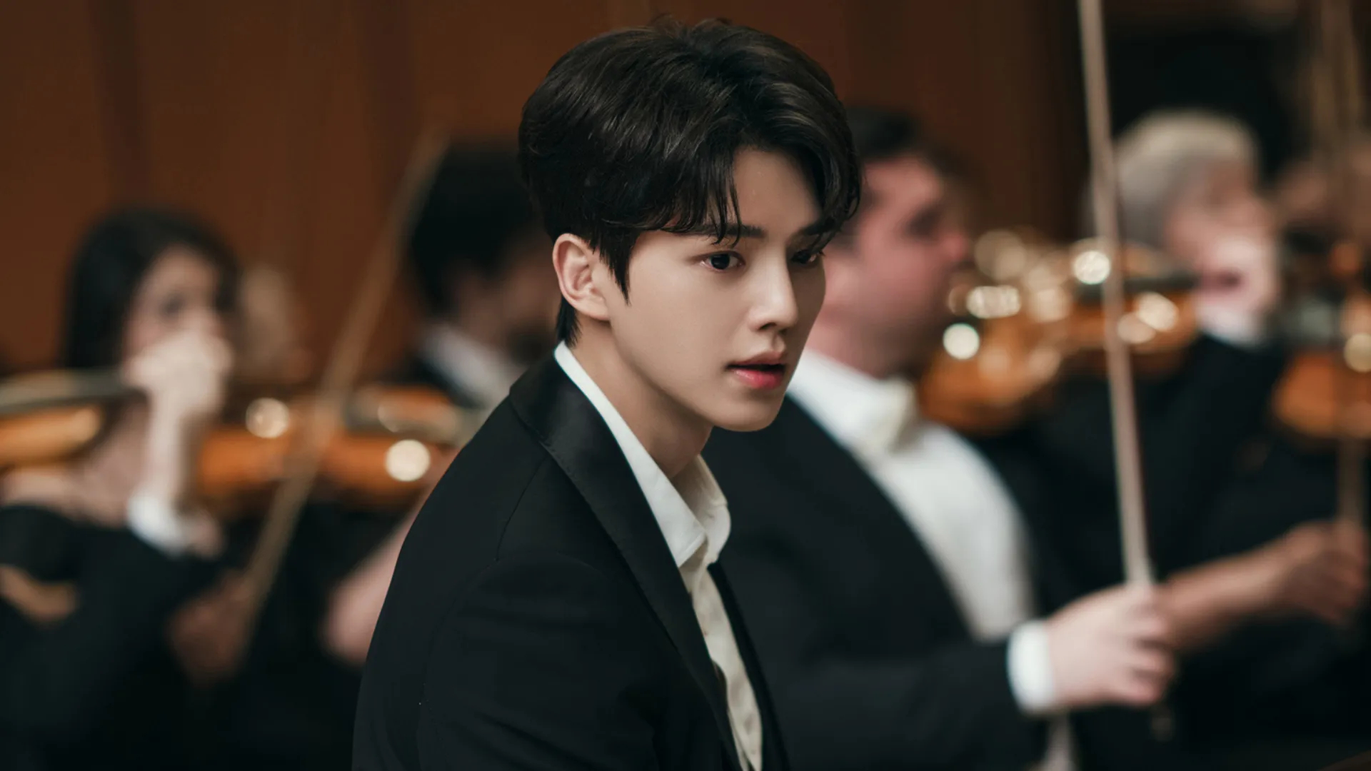 A Korean man (actor Song Kang) wears a suit while sitting in front of a blurred orchestra, for the K-drama 'Four Hands, Two Sonatas.'