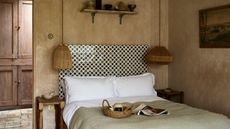 Image of a bedroom painted in a dusty pink limewash. There is a bed with white and sage green bedding and a tiled headboard made with black and white tiles. There are two rattan pendant lights hanging over wooden side tables on either side of the bed.