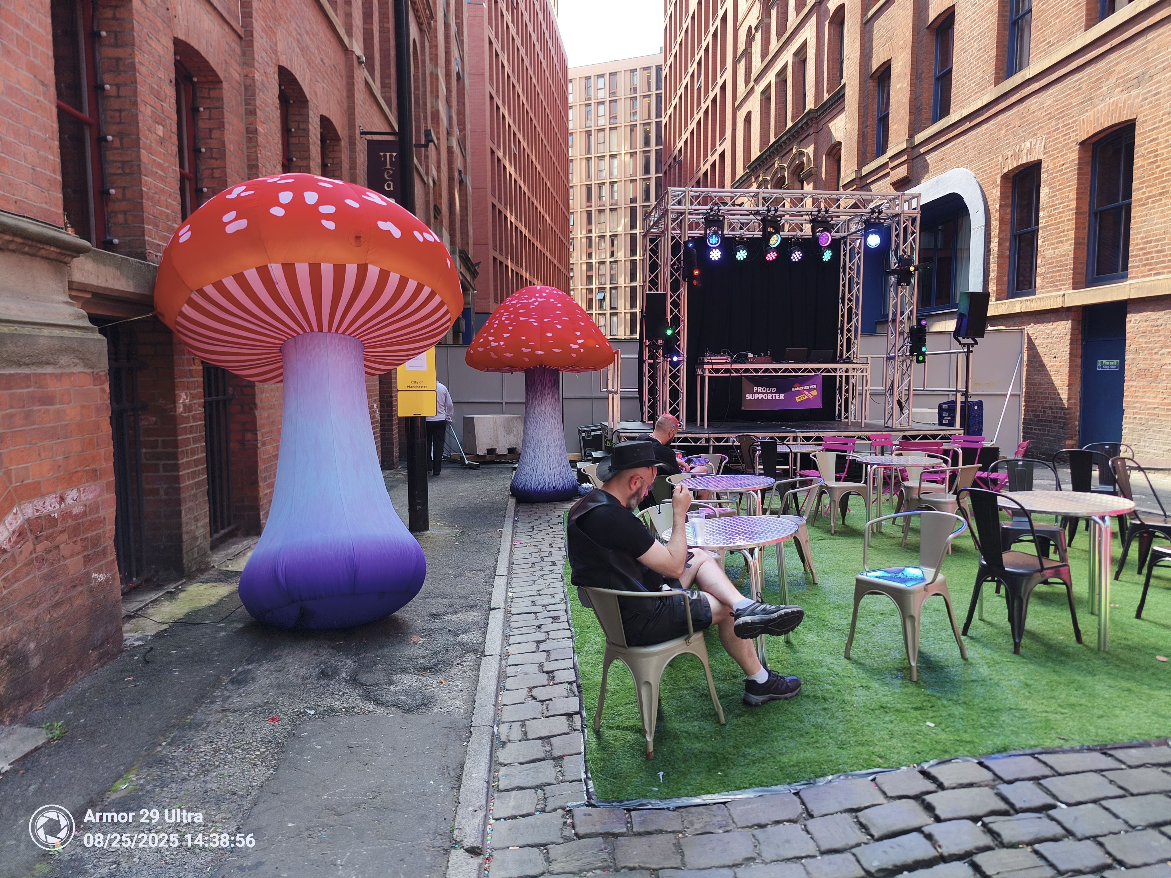 A small outdoor seating area with a stage is set up between two brick buildings, featuring two large, inflatable mushrooms and a person sitting at a table.