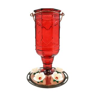 A glass red bottle hummingbird feeder with a copper base with white flowers adorning it