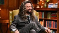 Mark Ballas, wearing a gray suit with dark tie, sits in a beige chair in a room of Alan Cumming's castle with bookshelves in the background on The Traitors Season 4.