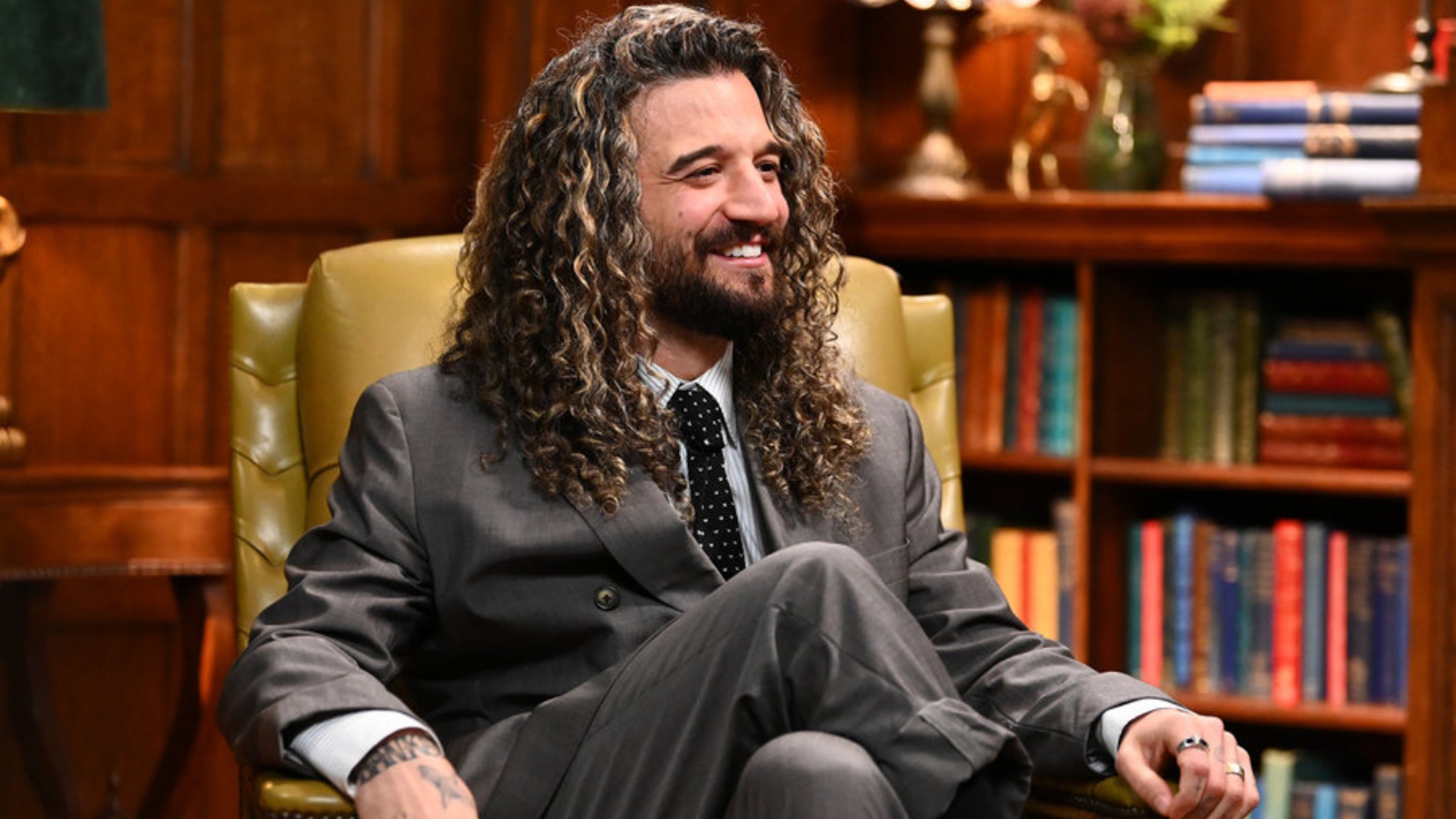 Mark Ballas, wearing a gray suit with dark tie, sits in a beige chair in a room of Alan Cumming's castle with bookshelves in the background on The Traitors Season 4.