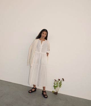 View of a female model wearing a long white dress with sleeves, flat black sandals and a light coloured cardigan which is sitting on her shoulders. The model has long hair with a middle part and she is standing in a space with white walls, grey floors and a plant