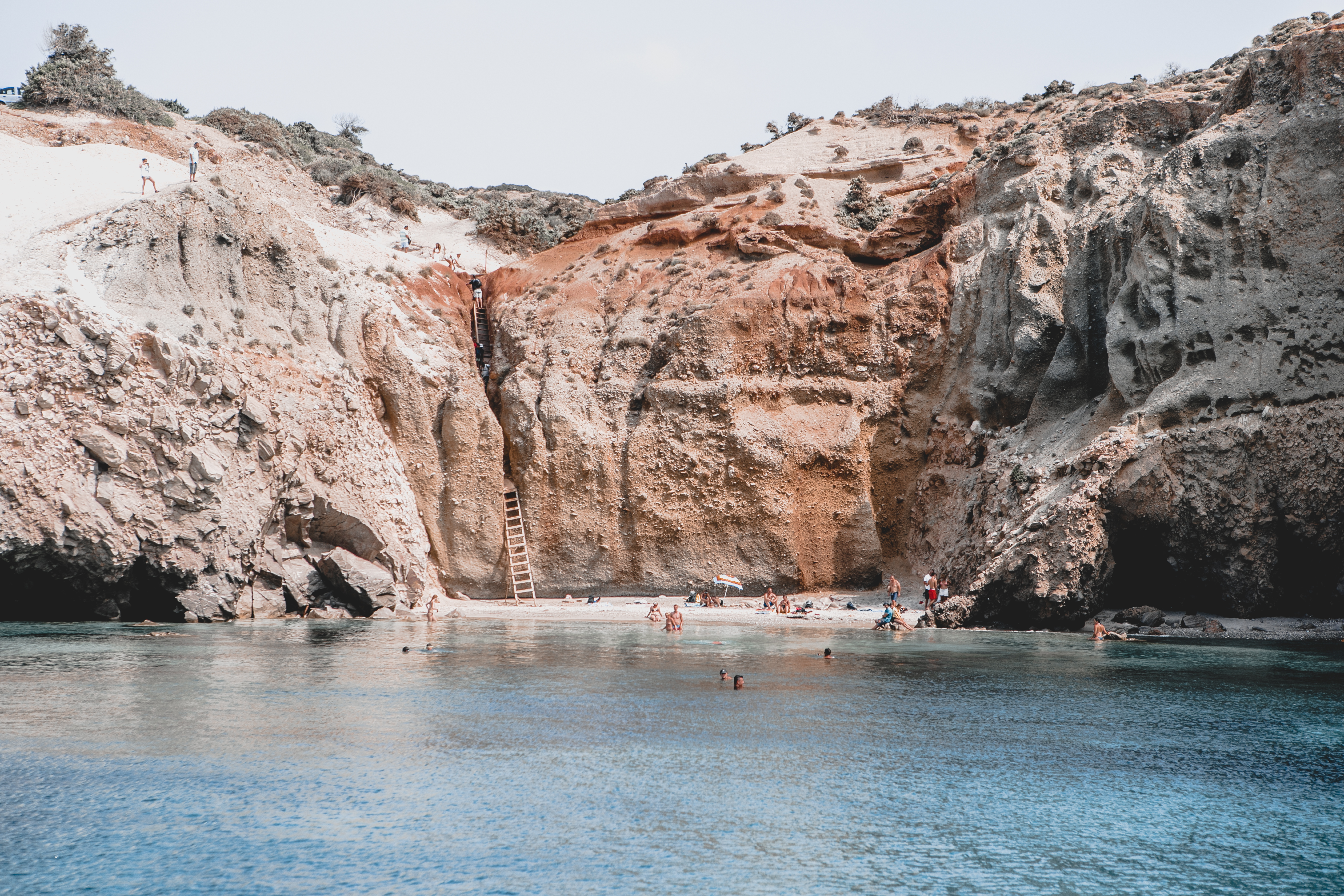 People swimming off a beach in Greece, backed by steep cliffs and accessible only via a wooden ladder