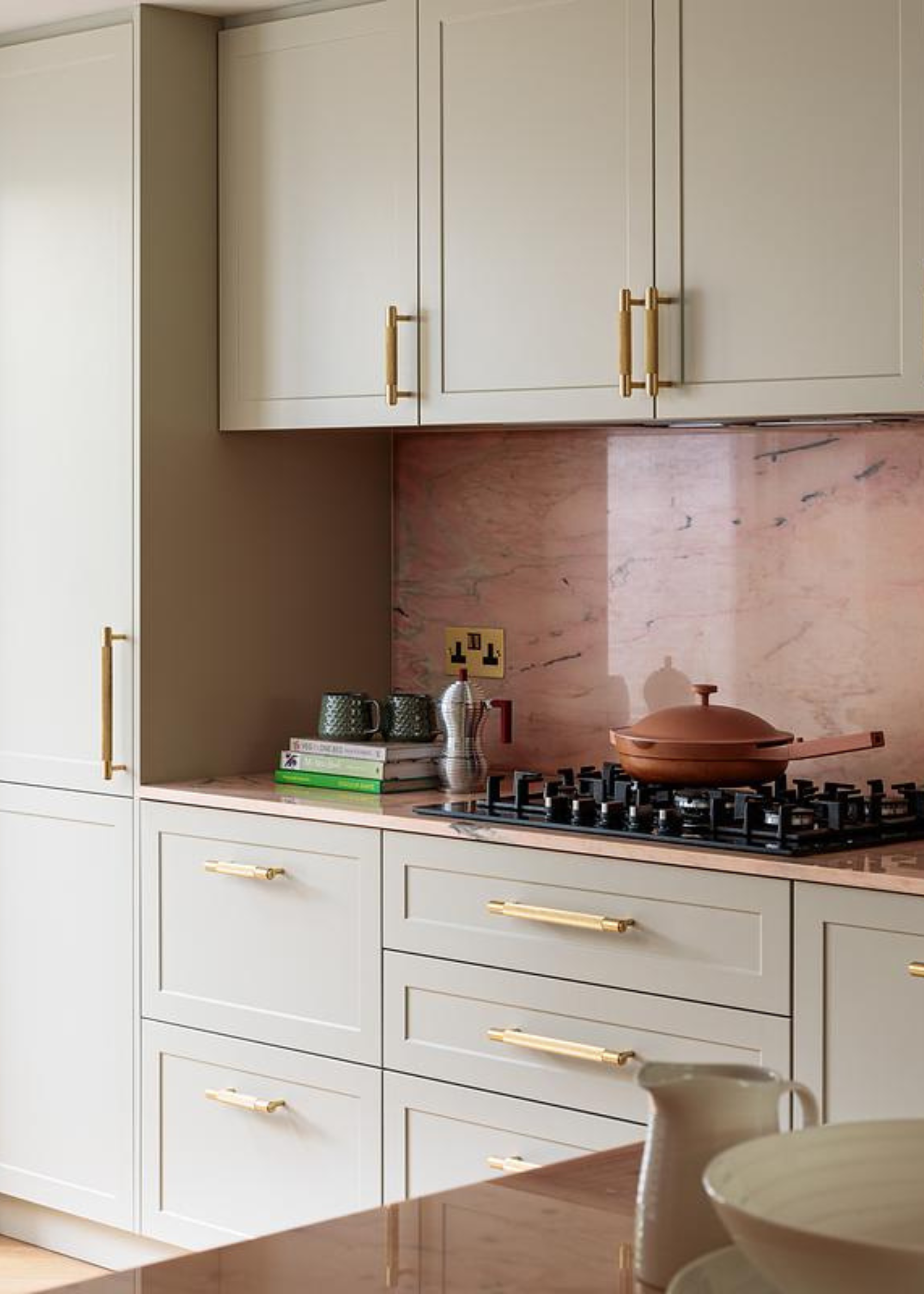 A kitchen with white cabinets and gold hardware, a gas stove with a terracotta-colored pan with a lid, an Alessi coffee pot and a stack of cook books with a pair of mugs