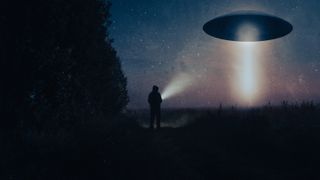 a flying saucer in the air on the top right shines a beam down into a dark overgrwoth. the silhouette of a person stands center on the dark ground, near a dark wall of trees on the left. they hold a flashlight toward the flying saucer.