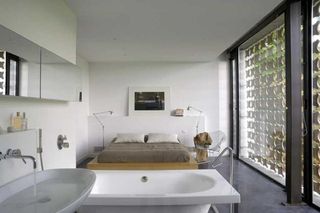 Bedroom, white walls, grey gloss floor, double bed on wooden platform, brown covers and white pillows, free stading white bath, white sink with running tab and filled bowl of water, black framed glass floor to ceiling windows, floor standing bedside lamps, mirror, wall art