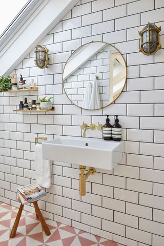 White metro tiling in bathroom with pink and white floor tiling, industrial wall sconces and gold faucets/pipes