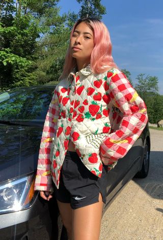 quilted patchwork jacket trend is shown on a woman standing by a car wearing a strawberry print jacket with basketball shorts