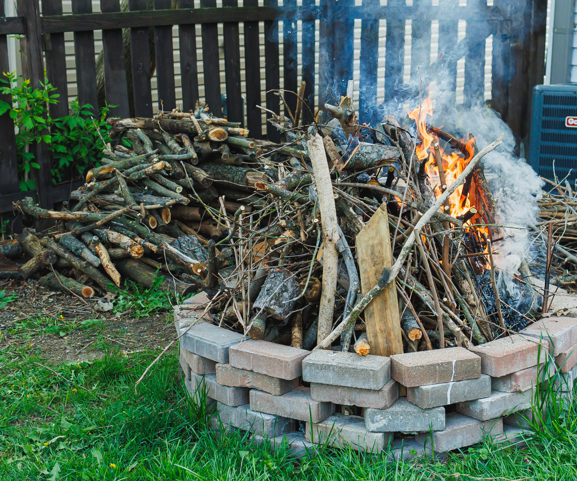 Circular bonfire with bricks stacked around the edges and sticks in the middle and a pile of logs in the background by the fence