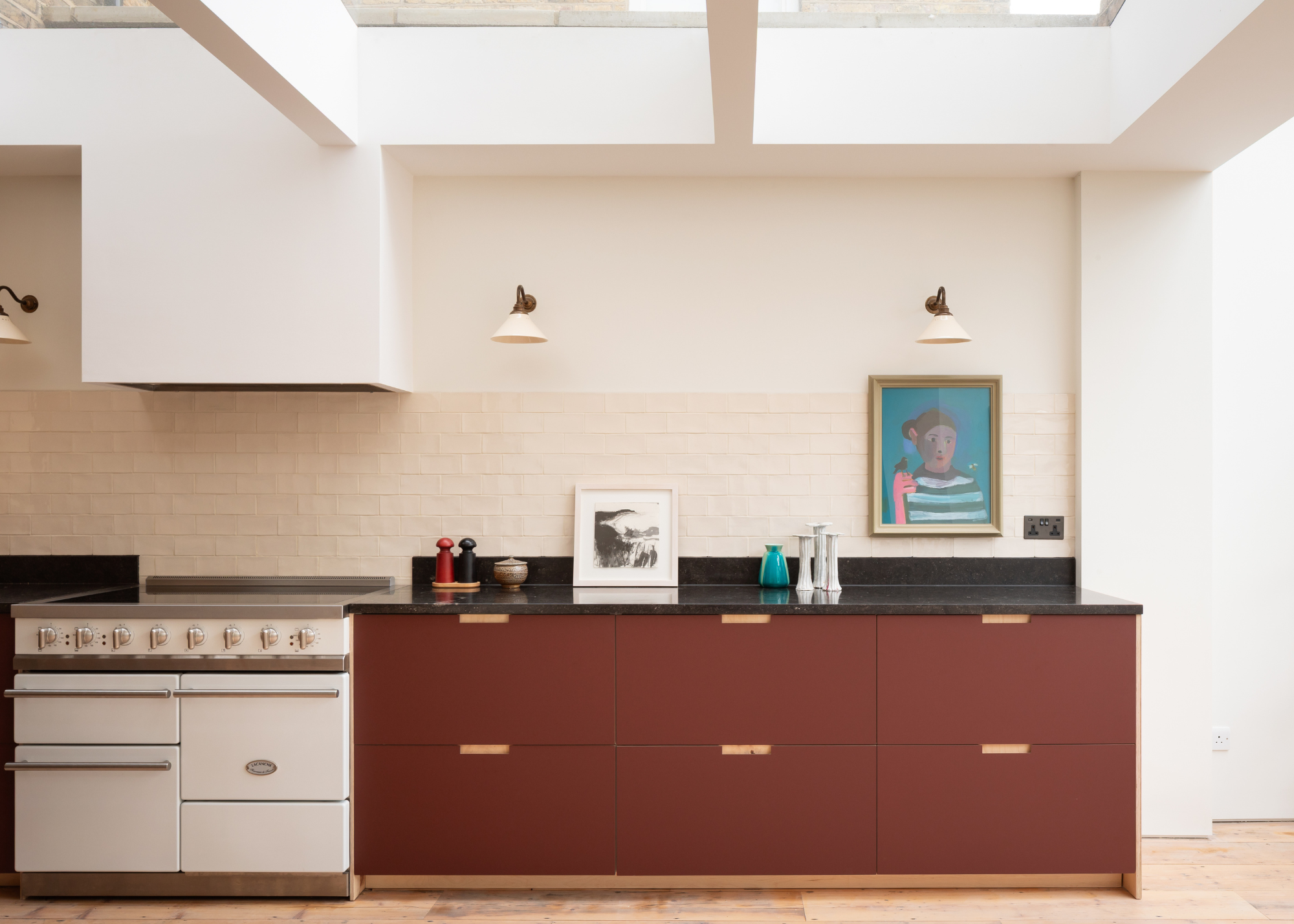 A bright kitchen with a white tile backsplash, maroon-brown kitchen drawers, and fluted wall sconces below skylights