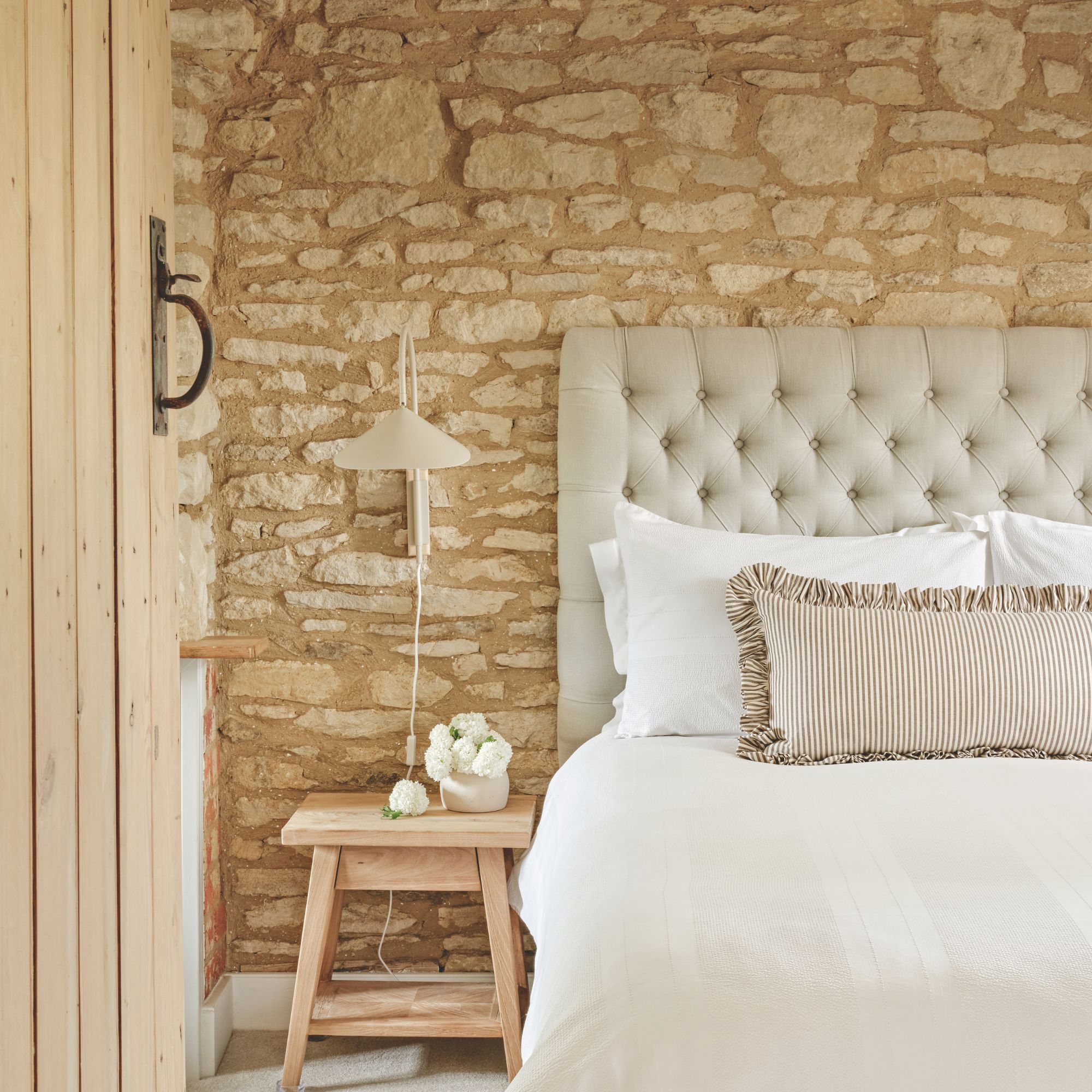 Cottage guest bedroom with stone wall and wooden door. Grey buttoned headboard with white bedding and striped frill-edge cushion and small wooden bedside table