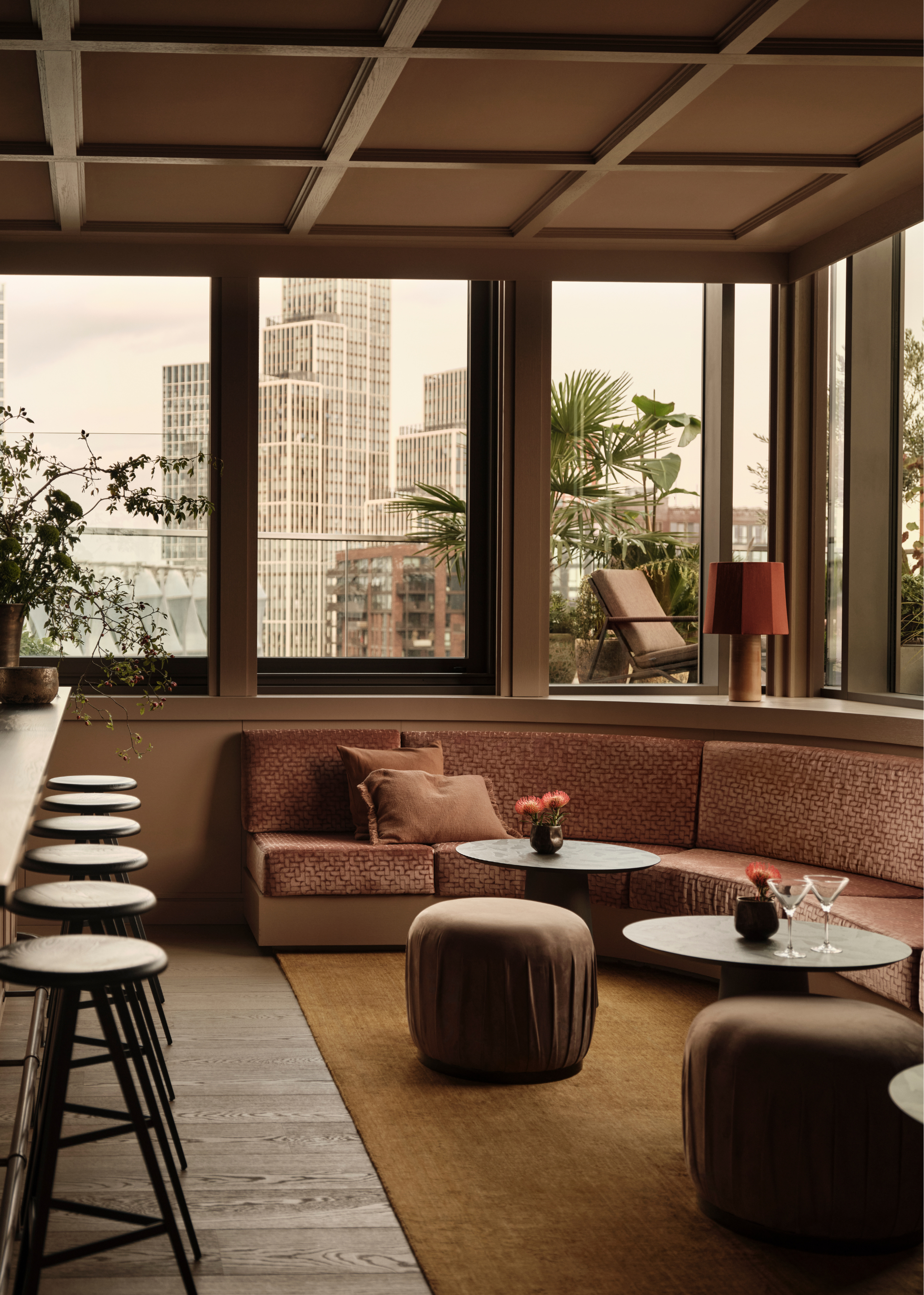 A modern open plan living space with curved banquette seating underneath large framed windows overlooking other skyrise apartments in London with a row of bar stools by the counter and two round coffee tables with poufs in front of the curved banquette