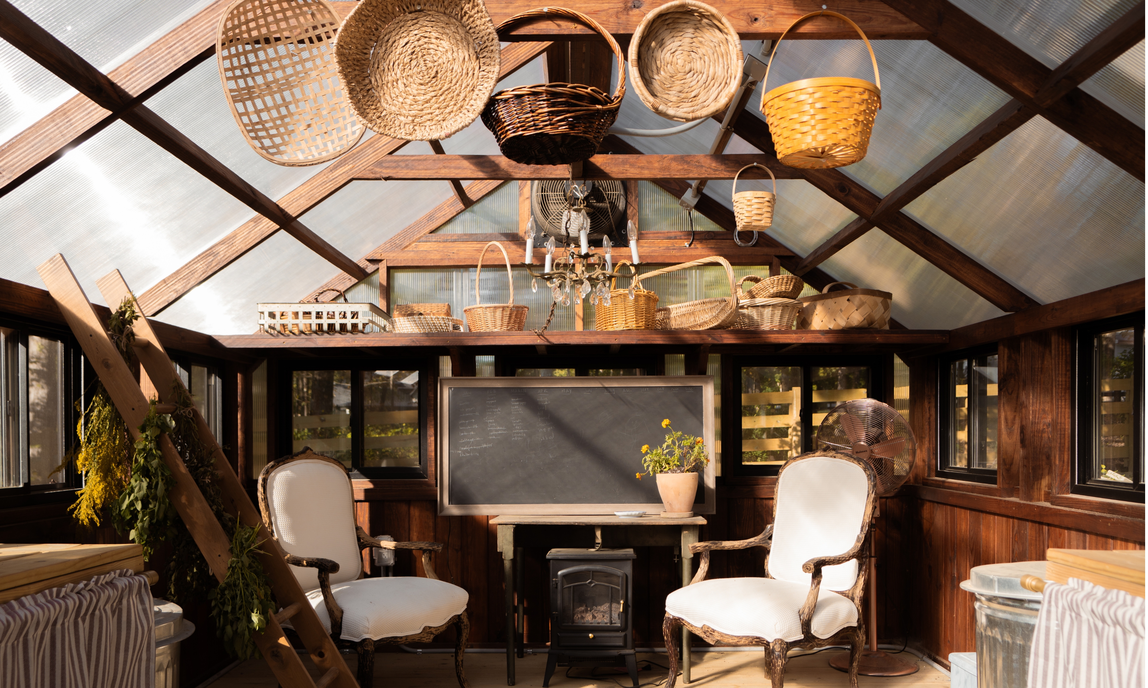 Greenhouse styled with antique chairs, a wood burning stove and wicker harvest baskets hanging from ceiling beam